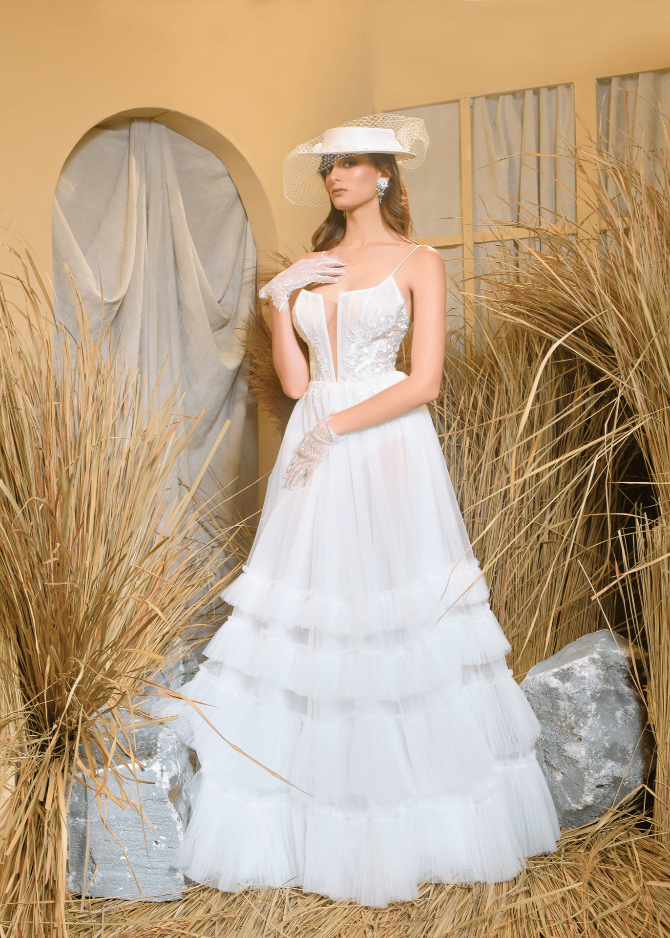 A woman standing in a traditional attire with a white floral wedding dress and a wide-brimmed hat, accessorized with a parasol, in a setting with a stone bench and dry plants in the background. Bridal gown available for stocking at One Fine Day Bridal Market New York