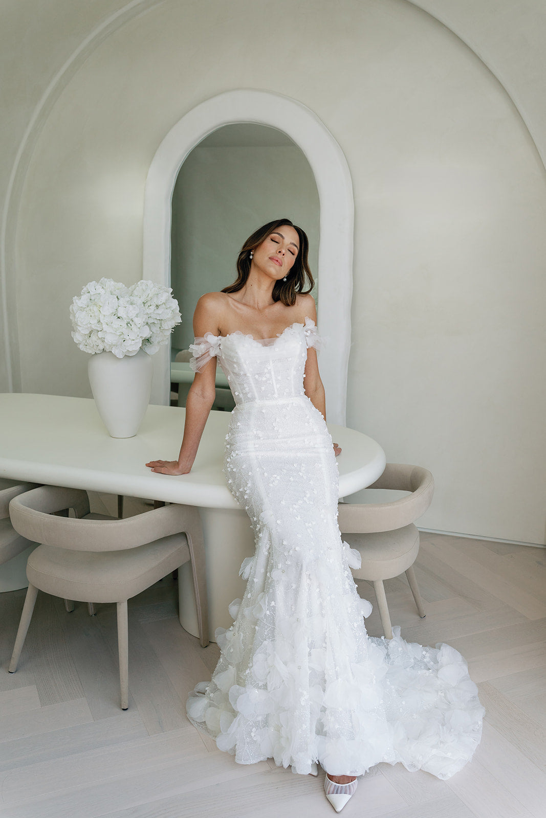 A woman standing by a round table in a elegant white bridal gown with floral embellishments and a high neck, with a large skirt for One Fine Day Bridal Market New York