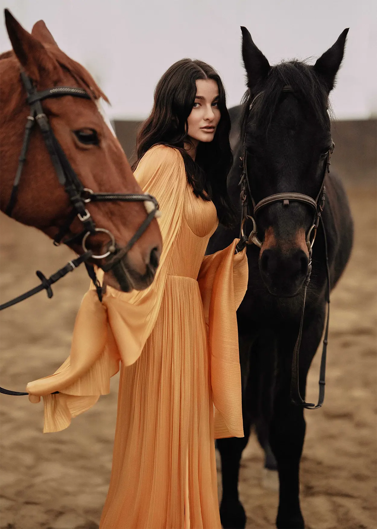 A woman wearing a yellow bridesmaid dress with long sleeves, standing between two horses in an outdoor setting.
