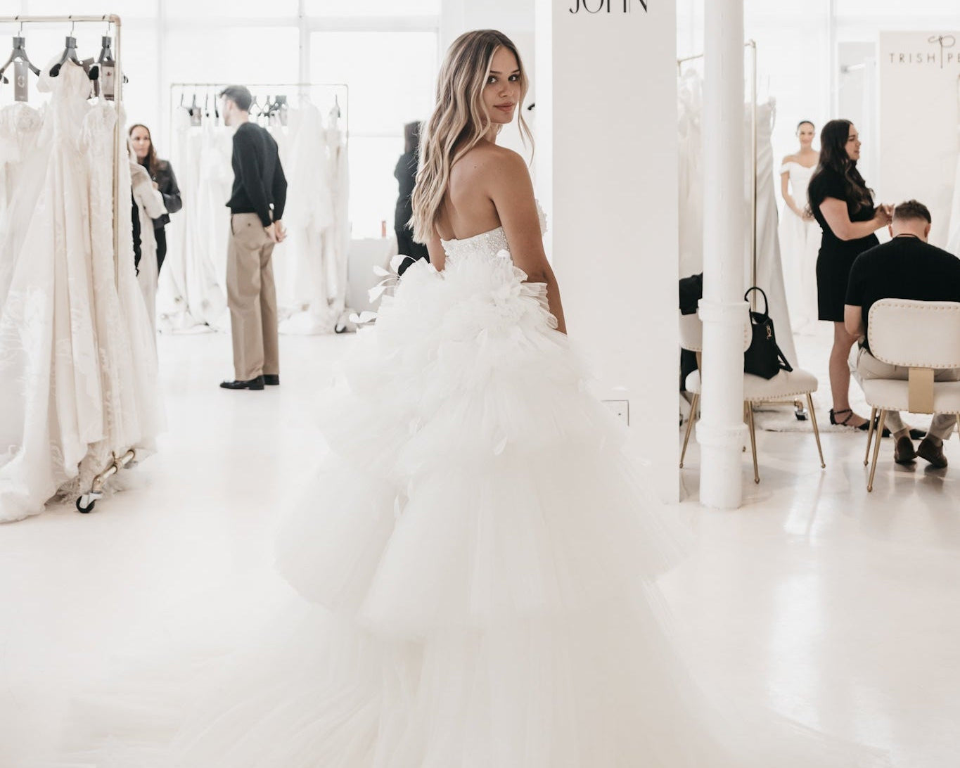 Woman in a white wedding dress standing in a bridal shop with 'Colby John' branding.
