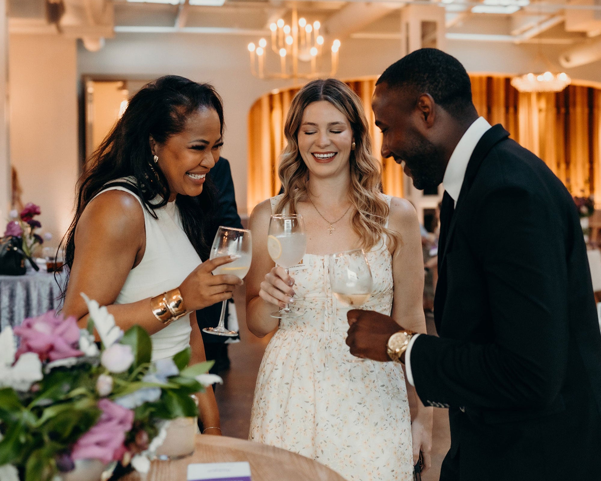Three people at the weddingpro lounge in the new york bridal market, two women and one man, smiling and holding drinks in a well-lit indoor setting.